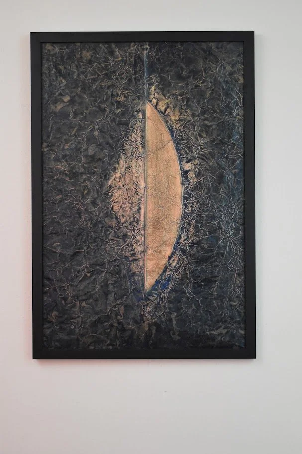 A golden crescent emerges from a dark, textured field. Displayed framed on a white wall.