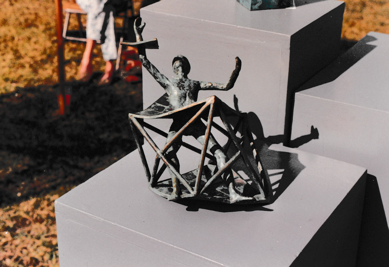 Small bronze figure seated within an angular geometric cage-like structure displayed on a pedestal outdoors. The figure raises one arm while the open framework creates strong triangular shapes around the body.