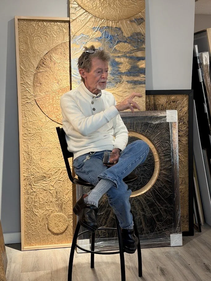 An older man with gray hair and beard, wearing a white sweater and blue jeans, is sitting cross-legged on a black stool, holding a smartphone. He is in front of abstract and textured artwork with gold and black elements, in an indoor setting.