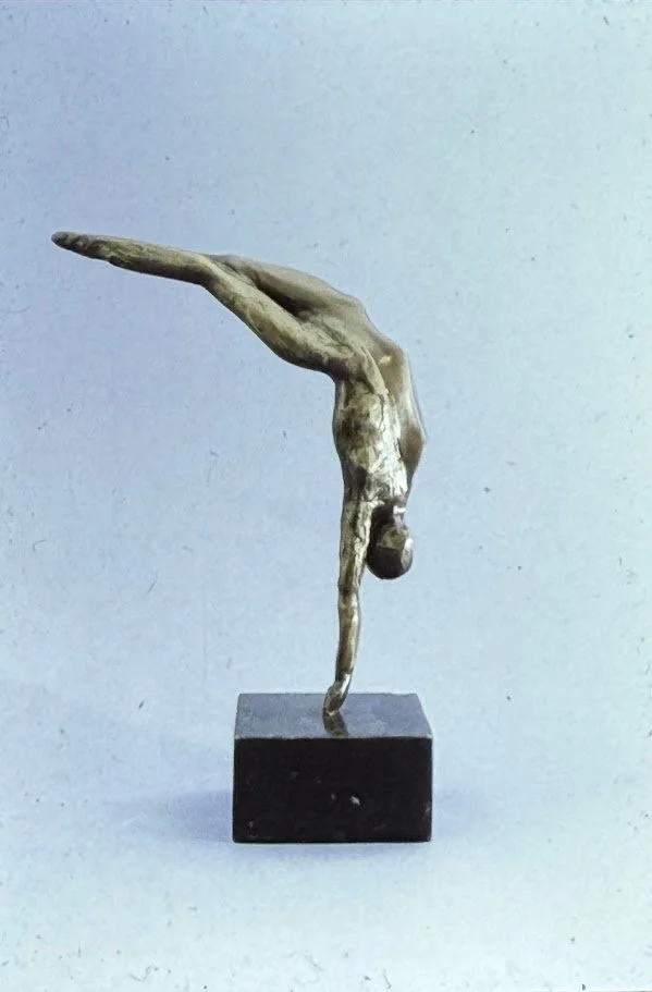 Minimal abstract bronze figure extending horizontally from a small pedestal, the elongated body suggesting a diver suspended in midair or a bird gliding through space.