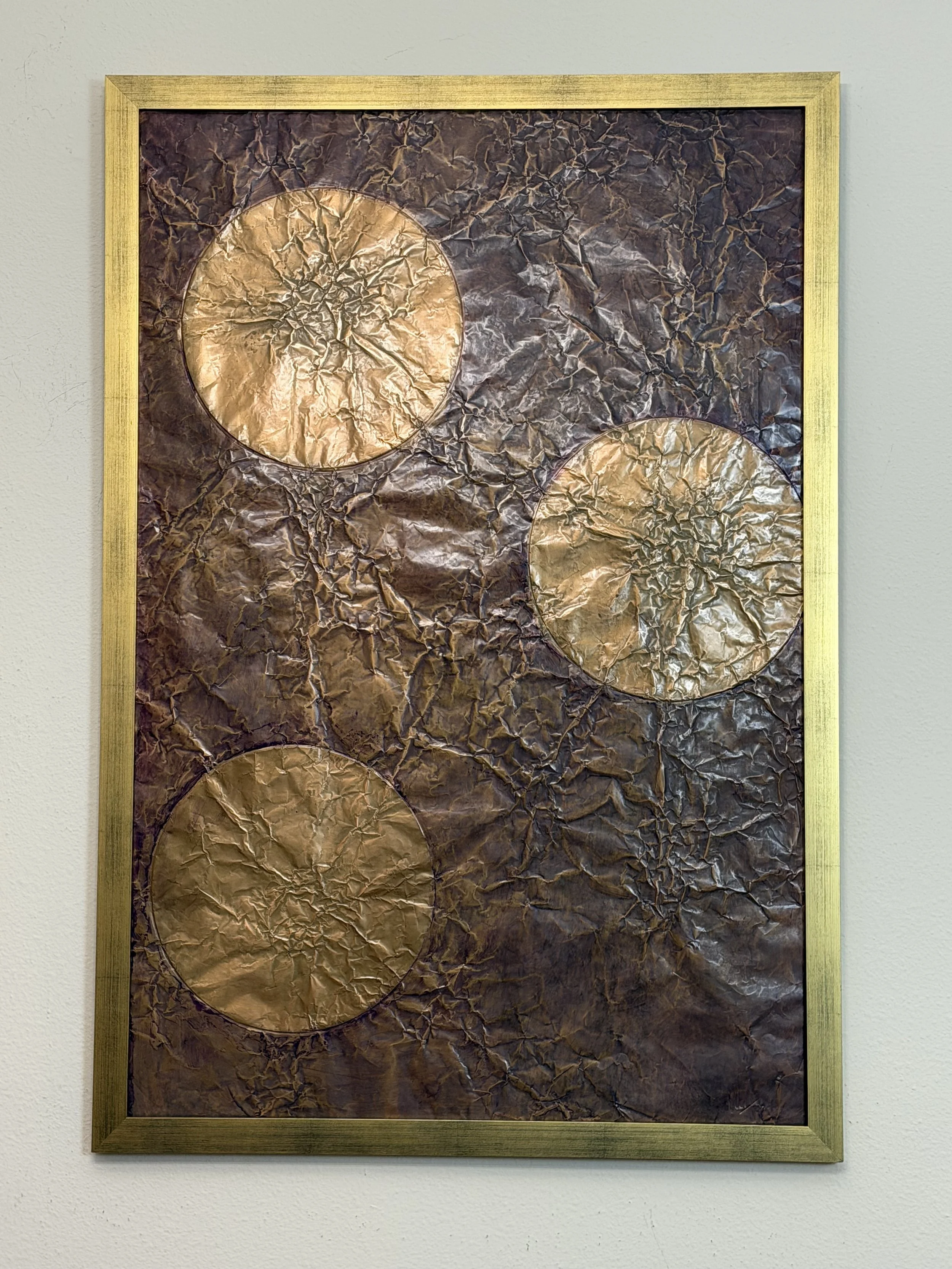 Three golden orbs float within a dark metallic field. Displayed framed on a white wall.