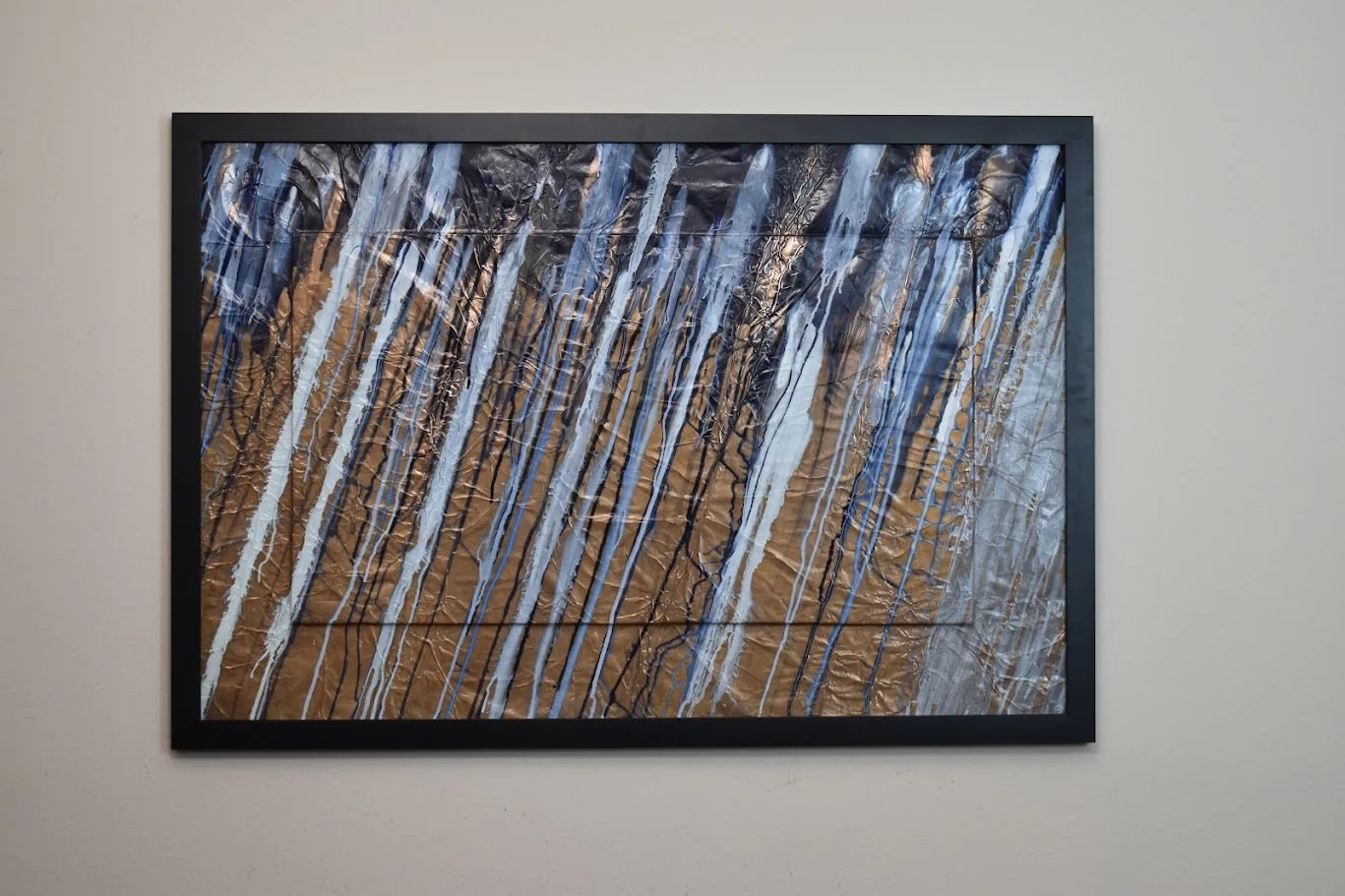 Diagonal blue, white, and black streaks sweep across a textured bronze field. Displayed framed on a white wall.
