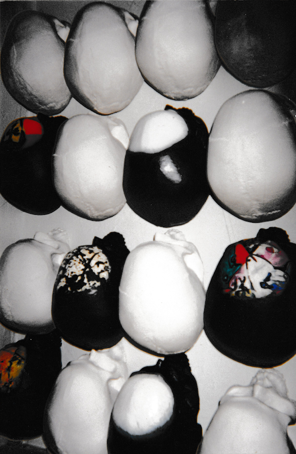 A collection of skull-shaped sculptures is displayed in a grid-like arrangement across a wall. Most of the forms are painted in black, white, or contrasting black-and-white with sections of color. 