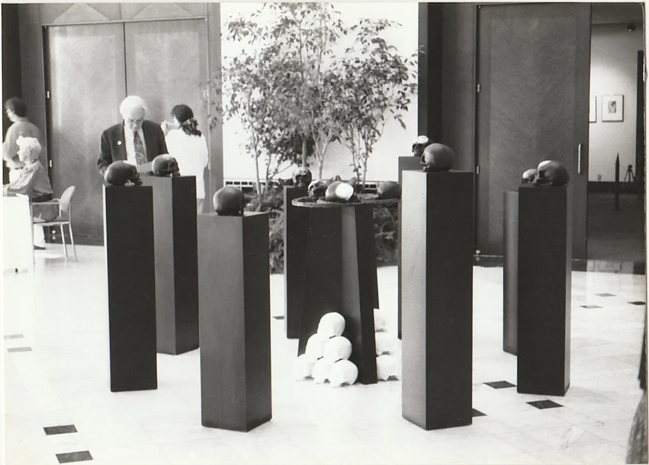 Several skull sculptures occupy a minimalist exhibition space, their dark surfaces contrasting against bright gallery walls and geometric pedestal forms.