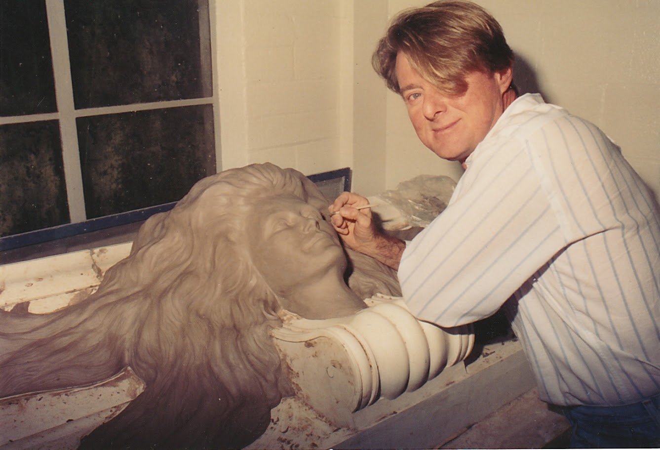 Whitney Wolf II sculpting a beautiful woman with long hair made out of clay. He is looking at the camera and smiling, and his hair is covering one of his eyes. EST 1990s