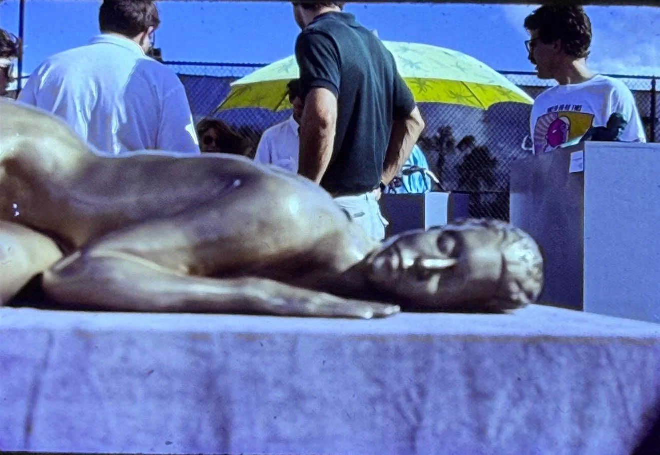 Closer view of the reclining bronze figure, focusing on the face and torso as spectators gather in the background.