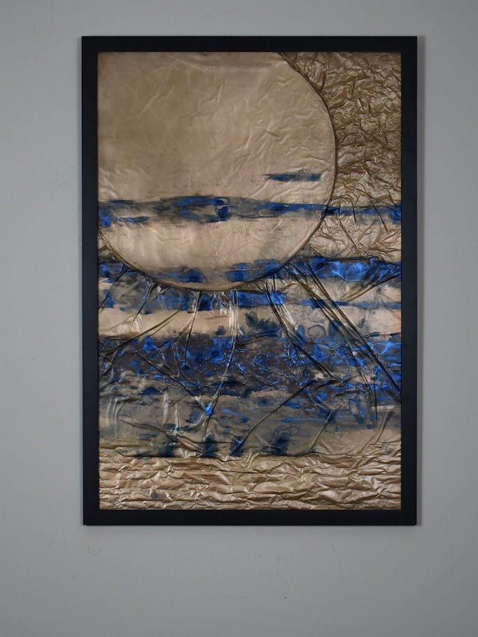 A pale planetary form emerges through layered blue, silver, and metallic currents. Displayed framed on a white wall.