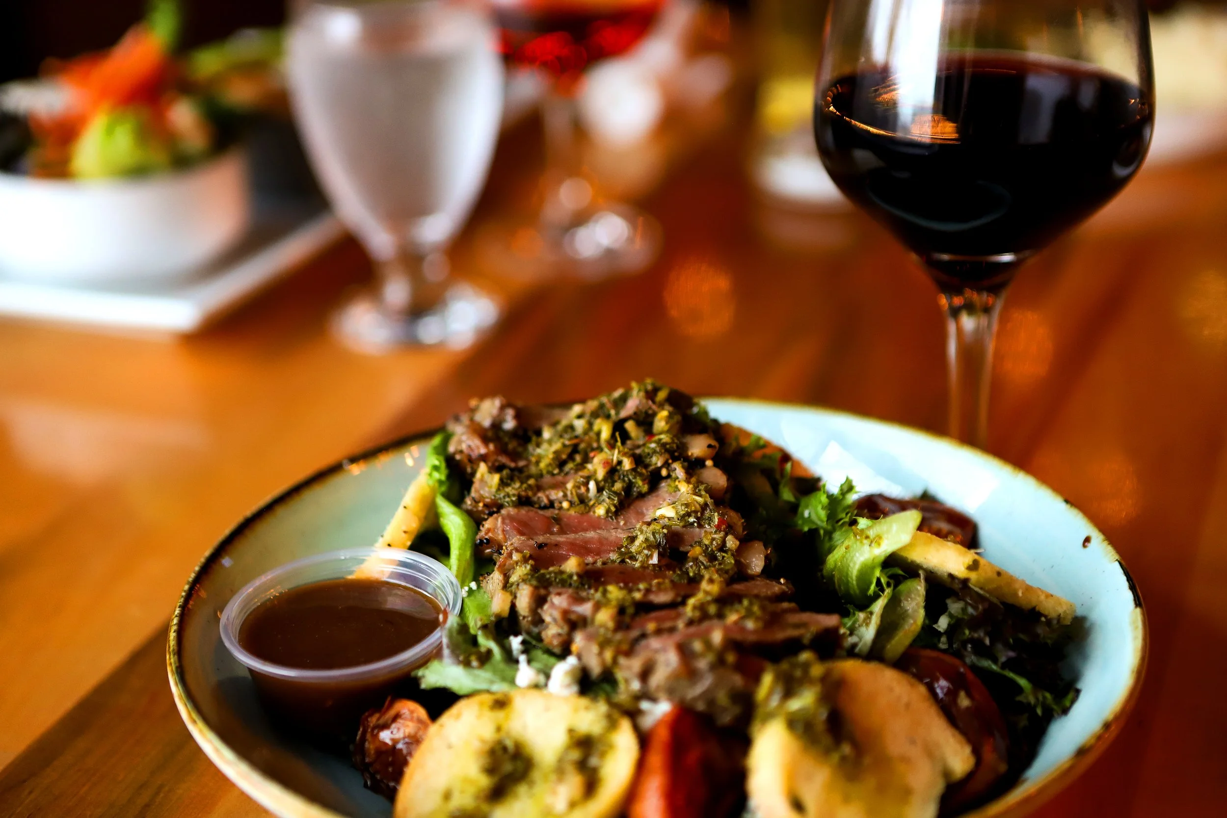 A bowl of sliced steak salad topped with herbs and sauce, with a small dish of sauce on the side, set on a wooden table with wine glasses and meals in the background.