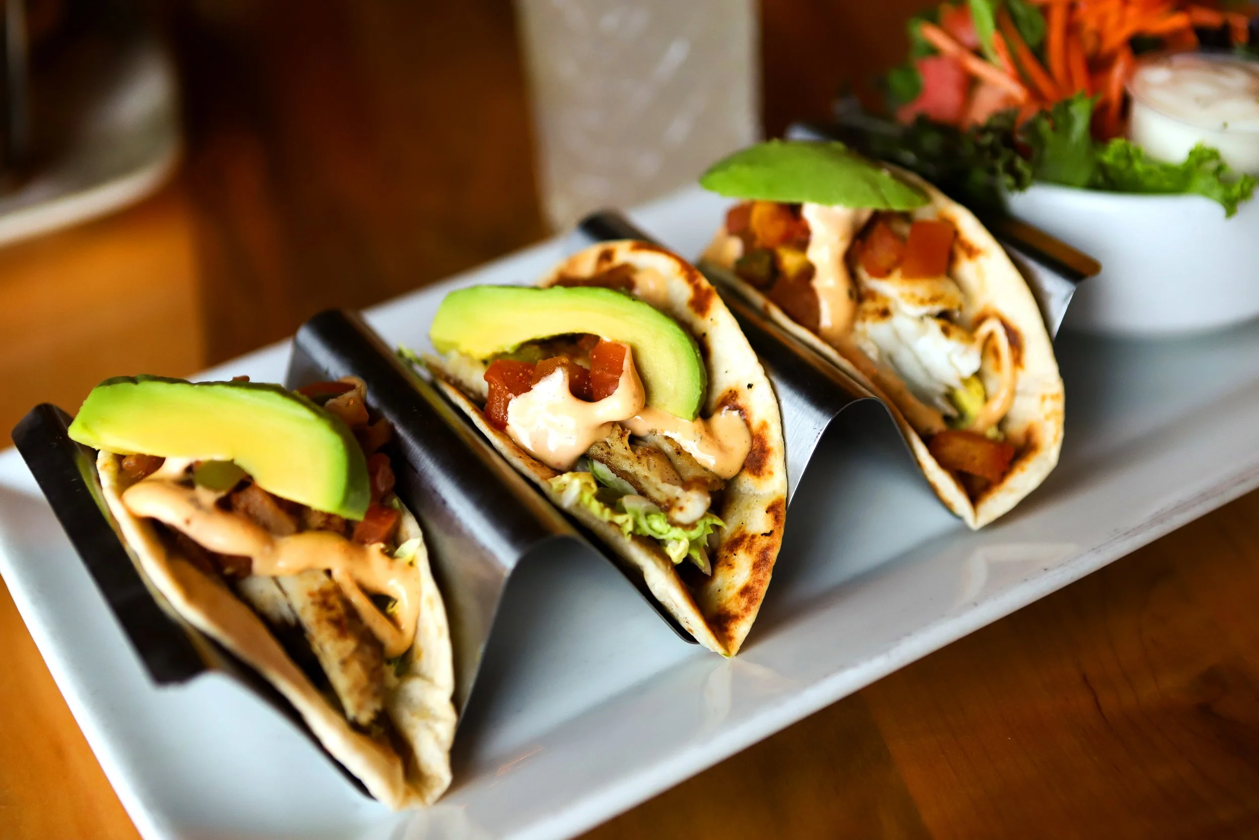 Three tacos filled with meat, avocado slices, lettuce, and sauce, served on a white plate with a side of greens and dipping sauce in a white bowl.