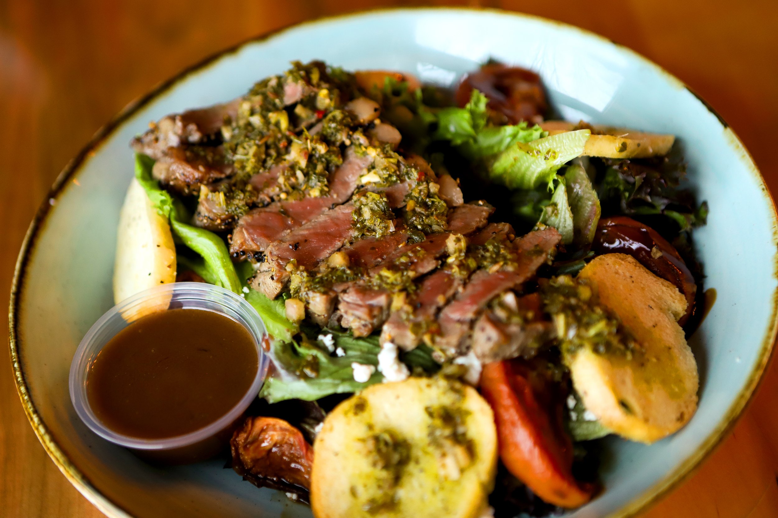 The Bruce Beef Salad includes sliced grilled steak topped with a green herb sauce, fresh green and purple lettuce, grilled vegetables, and a side of balsamic in a small cup.