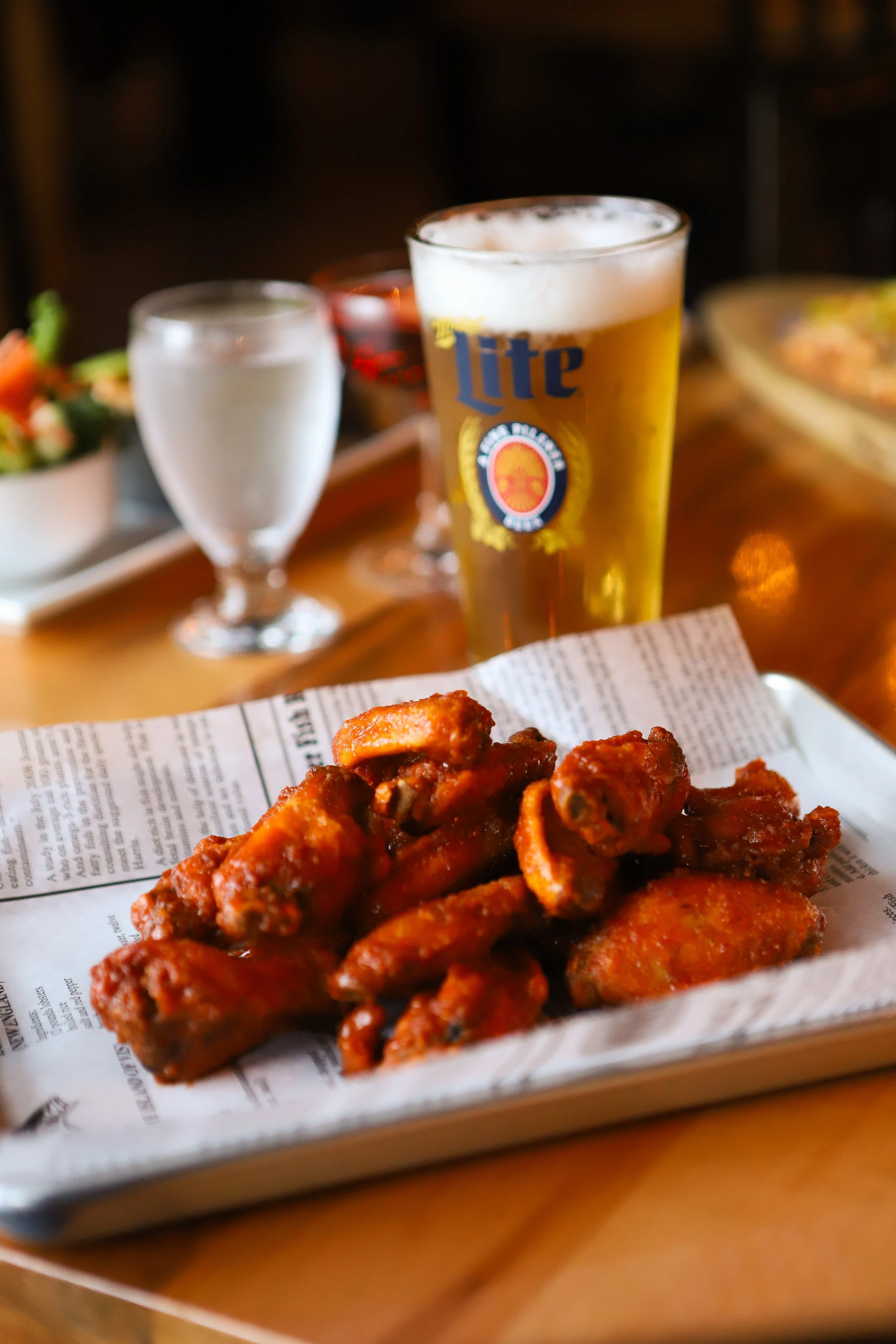 Chicken wings on a sheet of paper, with a background featuring a glass of beer, a glass of water, and some plates of food.