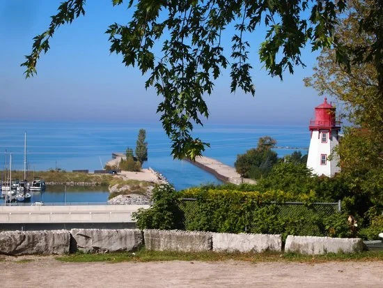 Kincardine lighthouse on Lake Huron, surrounded by trees and a stone barrier, with a view of the lake and a marina in the distance.
