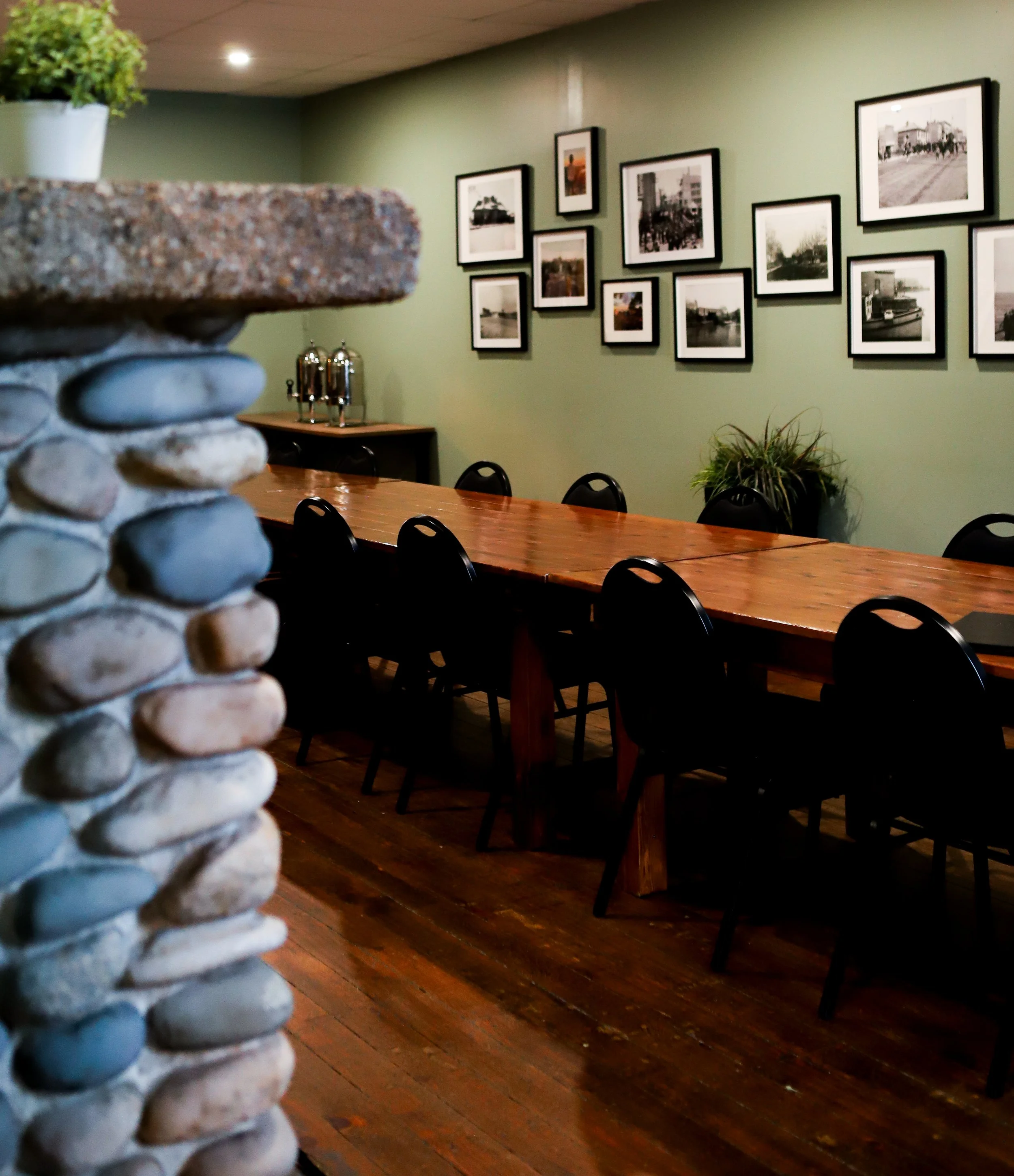 A fireplace with smooth stones in the foreground. In the background, a wooden table and black chairs are inside a meeting room with a green wall decorated with black-and-white photographs.