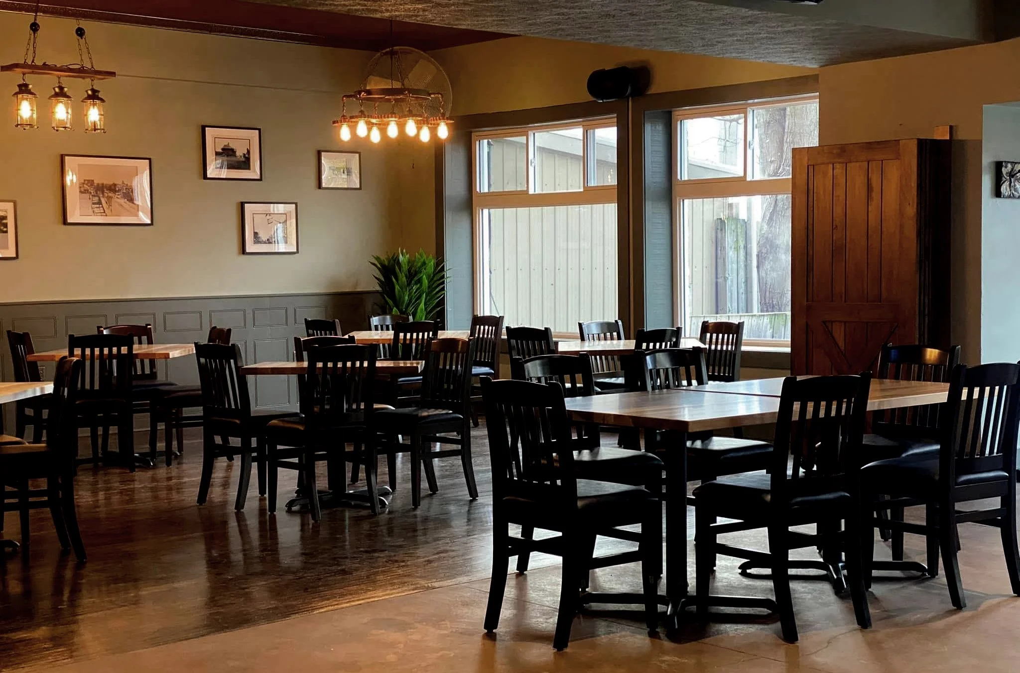 The Bruce Dining Room. Empty restaurant dining area with wooden tables and black chairs, large windows, green plant, framed pictures on the wall, and warm lighting fixtures.