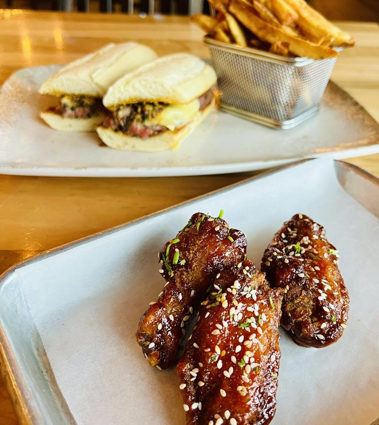 The Bruce Social Media. Close-up of glazed chicken wings topped with sesame seeds and chopped green herbs on a white tray, with a plate of breakfast sandwiches and fries in the background.
