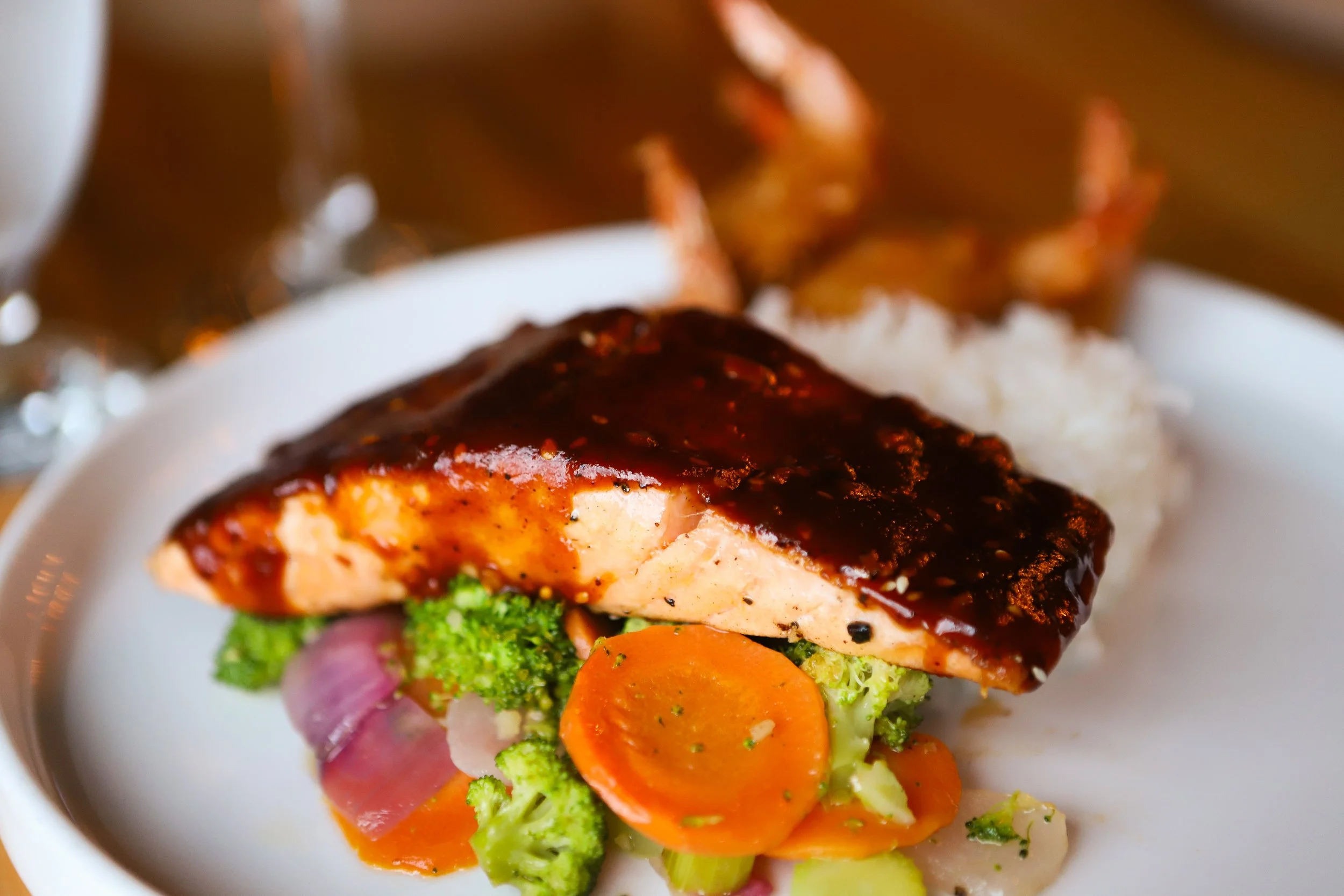 Close-up of a plate with a cooked piece of salmon topped with dark sauce, accompanied by steamed broccoli, sliced carrots and coconut shrimp.