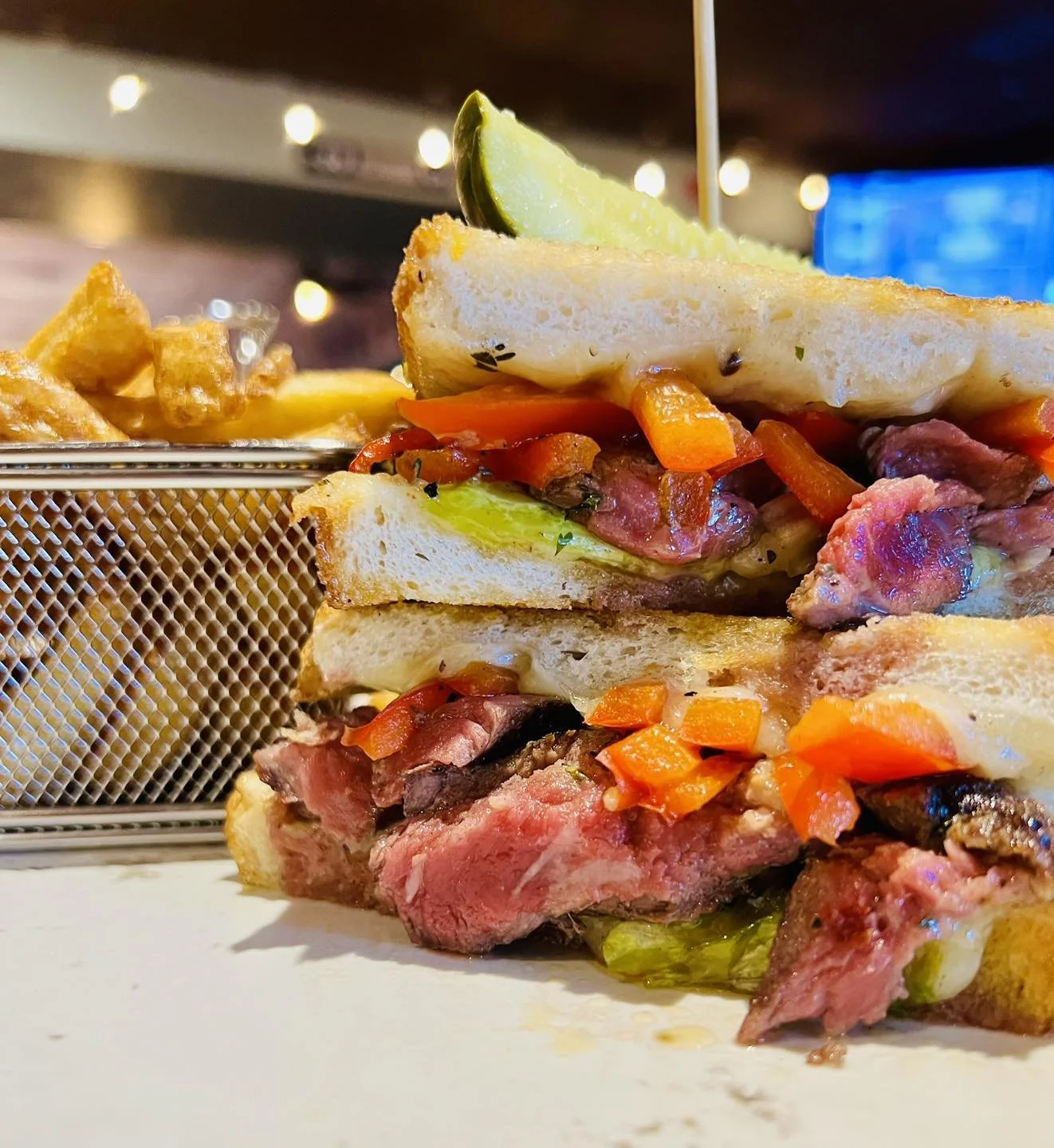 The Bruce Social Media. Close-up of a sandwich with roasted meats, vegetables, melted cheese, and a lime wedge on top, with a side of waffle fries in a wire basket in the background.