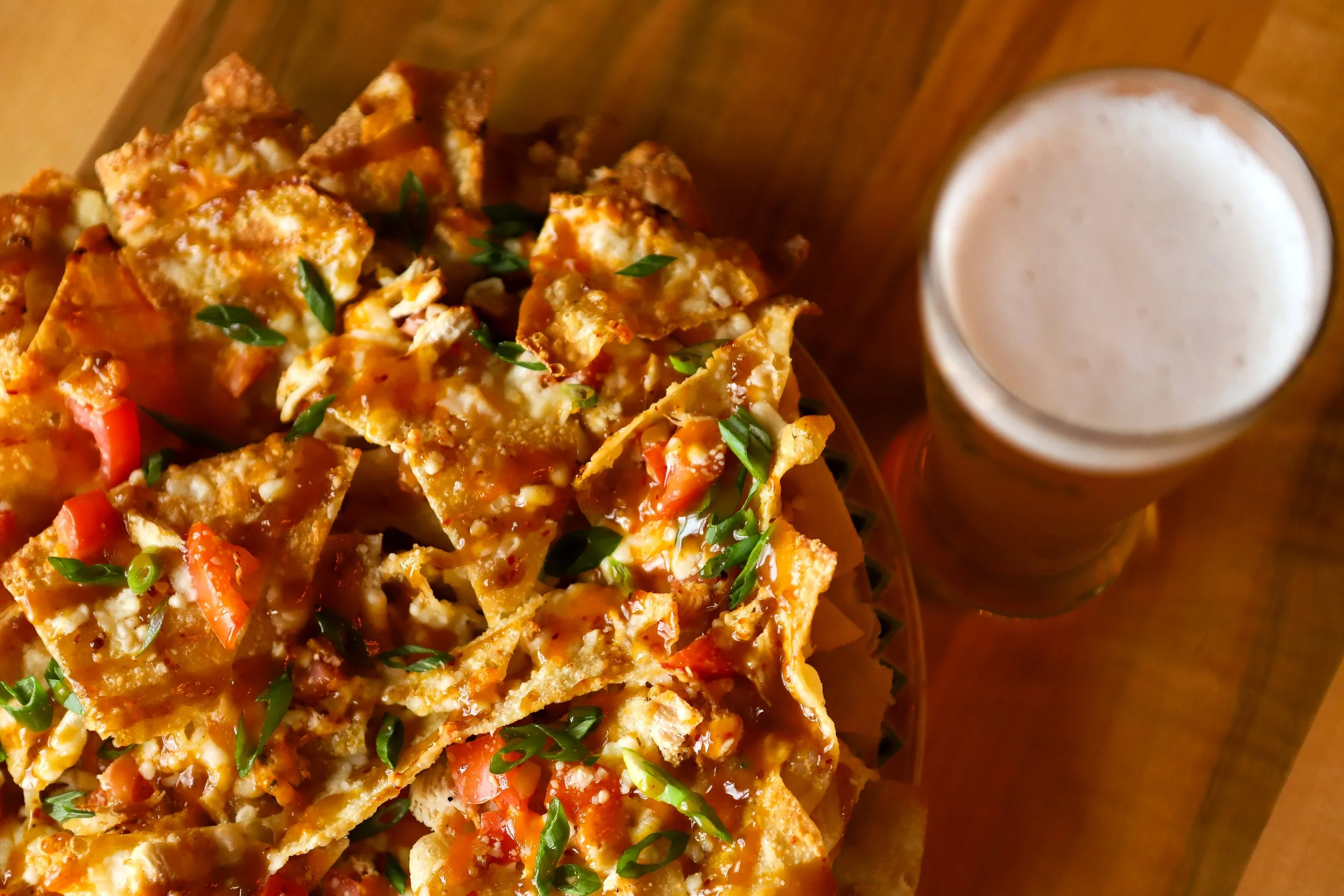 Close-up of nachos topped with cheese, diced tomatoes, and green herbs, with a glass of frothy beer nearby on a wooden table.