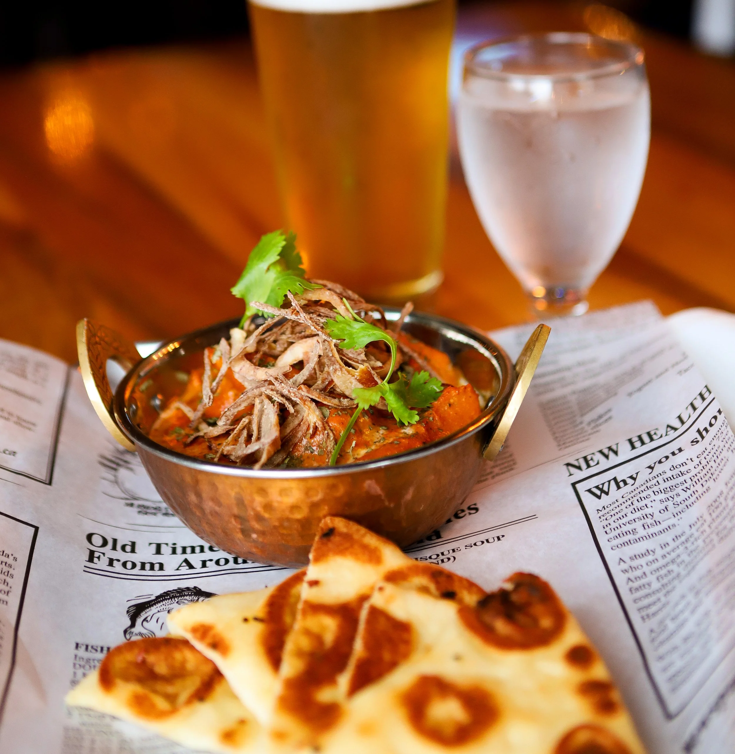 Close-up of a bowl of butter chicken garnished with cilantro, with beer, water, and sliced naan on a newspaper background.