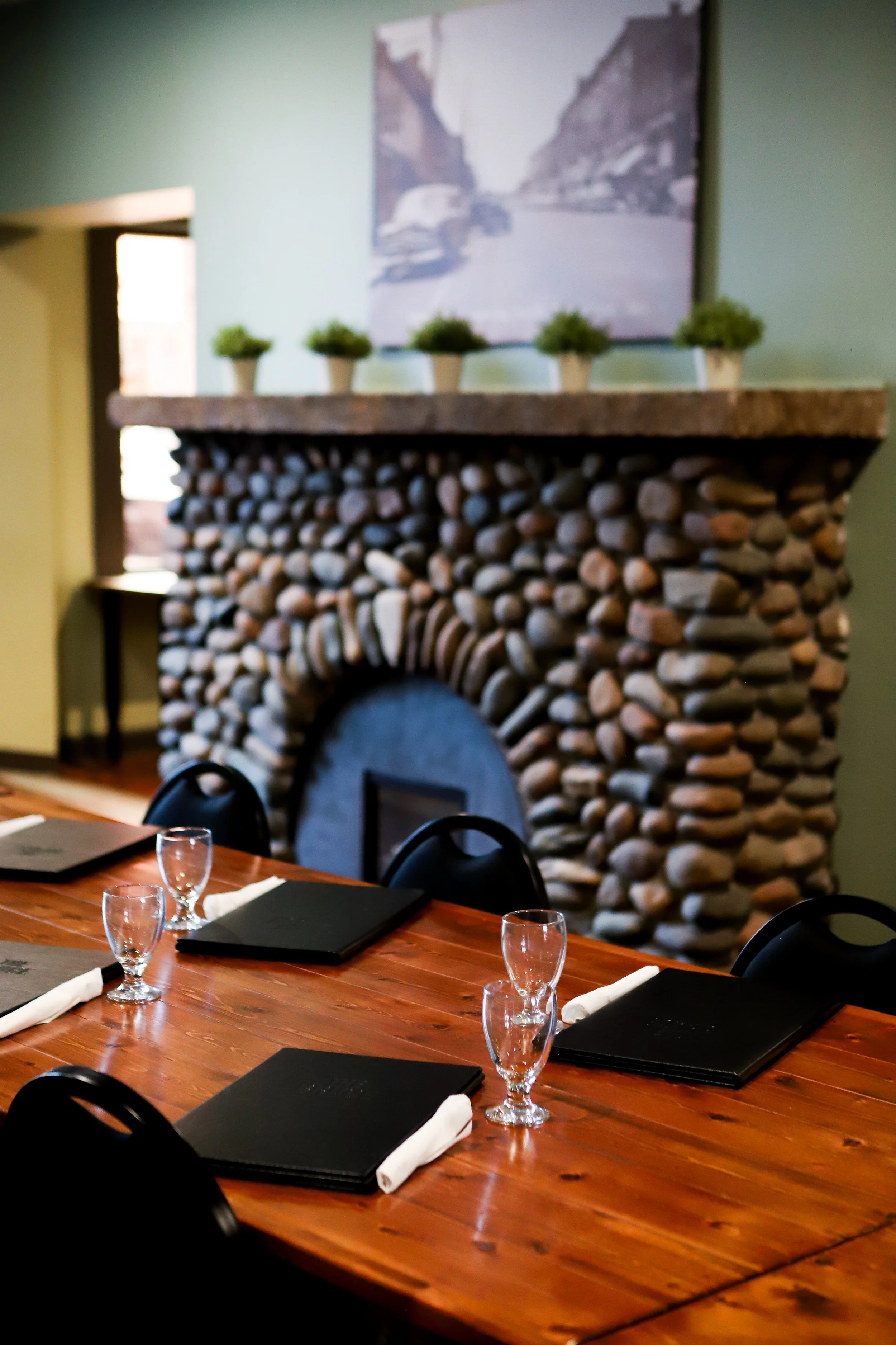The Mackenzie Room A dining table set with black menus, white napkins, and water glasses, in front of a stone fireplace with six small potted plants on top, and a painting hanging above it.