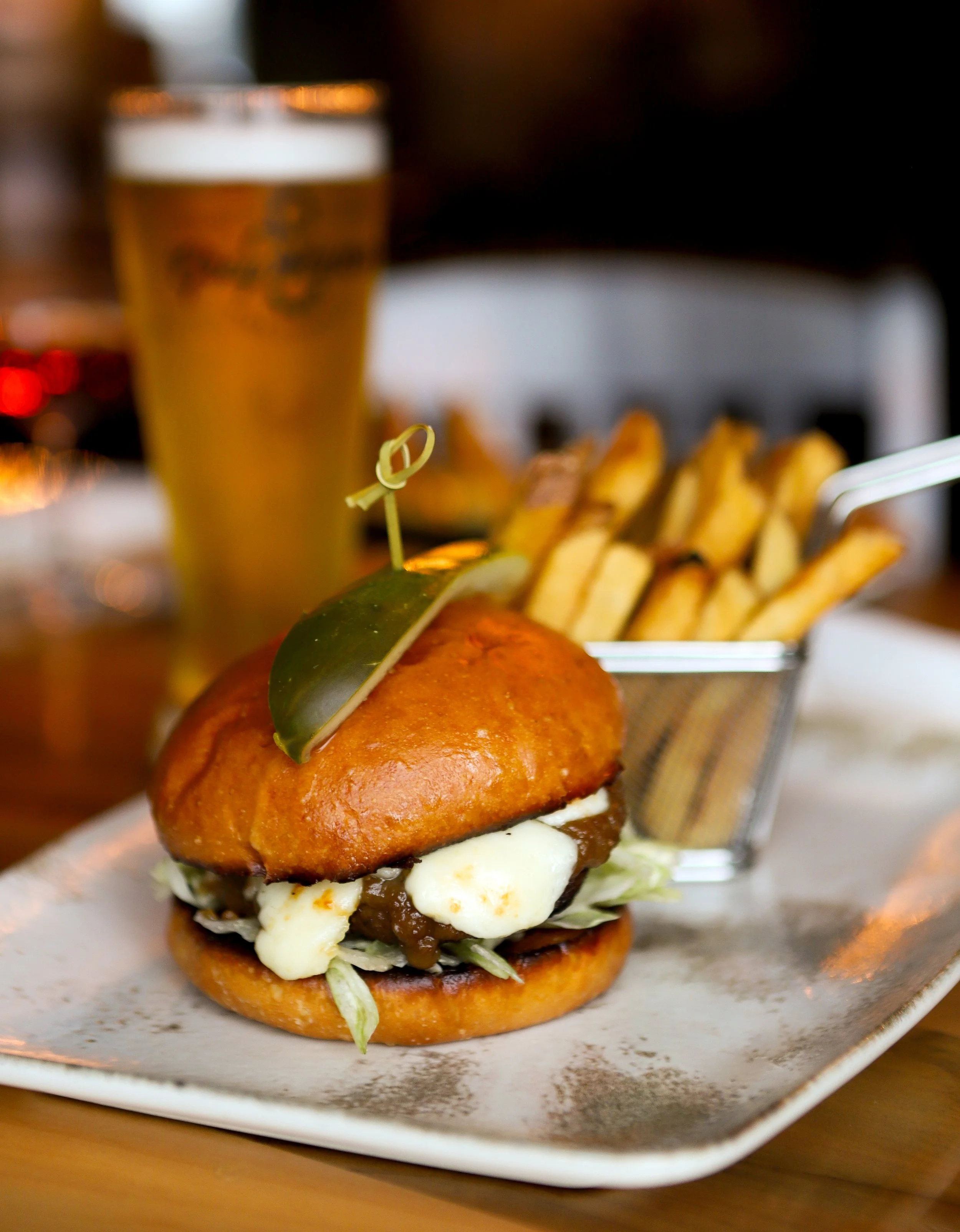 Close-up of a gourmet burger with lettuce, cheese, and sauce, served with a side of French fries and a glass of beer in the background.