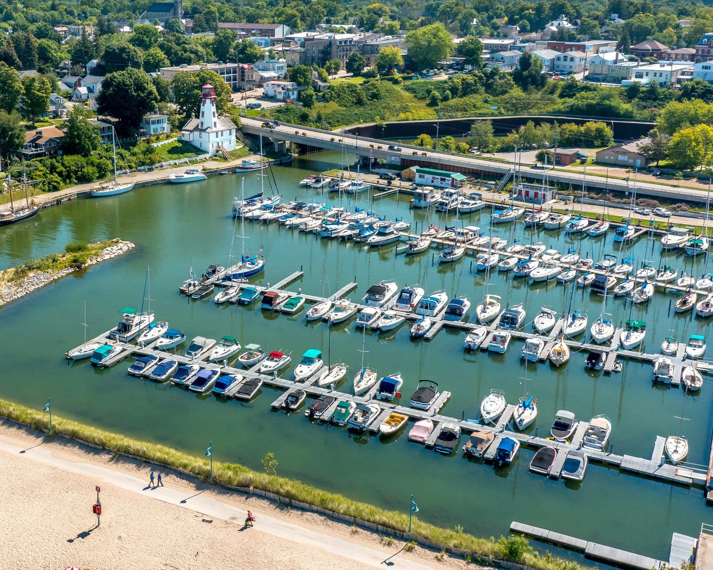An aerial view of a marina with numerous boats docked along the piers, a lighthouse, and a bridge crossing over the water. Surrounding the marina are green trees and a town with houses and buildings, including The Bruce restaurant.