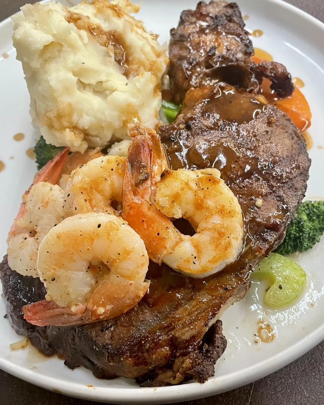 The Bruce Social Media. Plate of food with mashed potatoes topped with gravy, shrimp, steak with gravy, and cooked vegetables including broccoli and carrots.