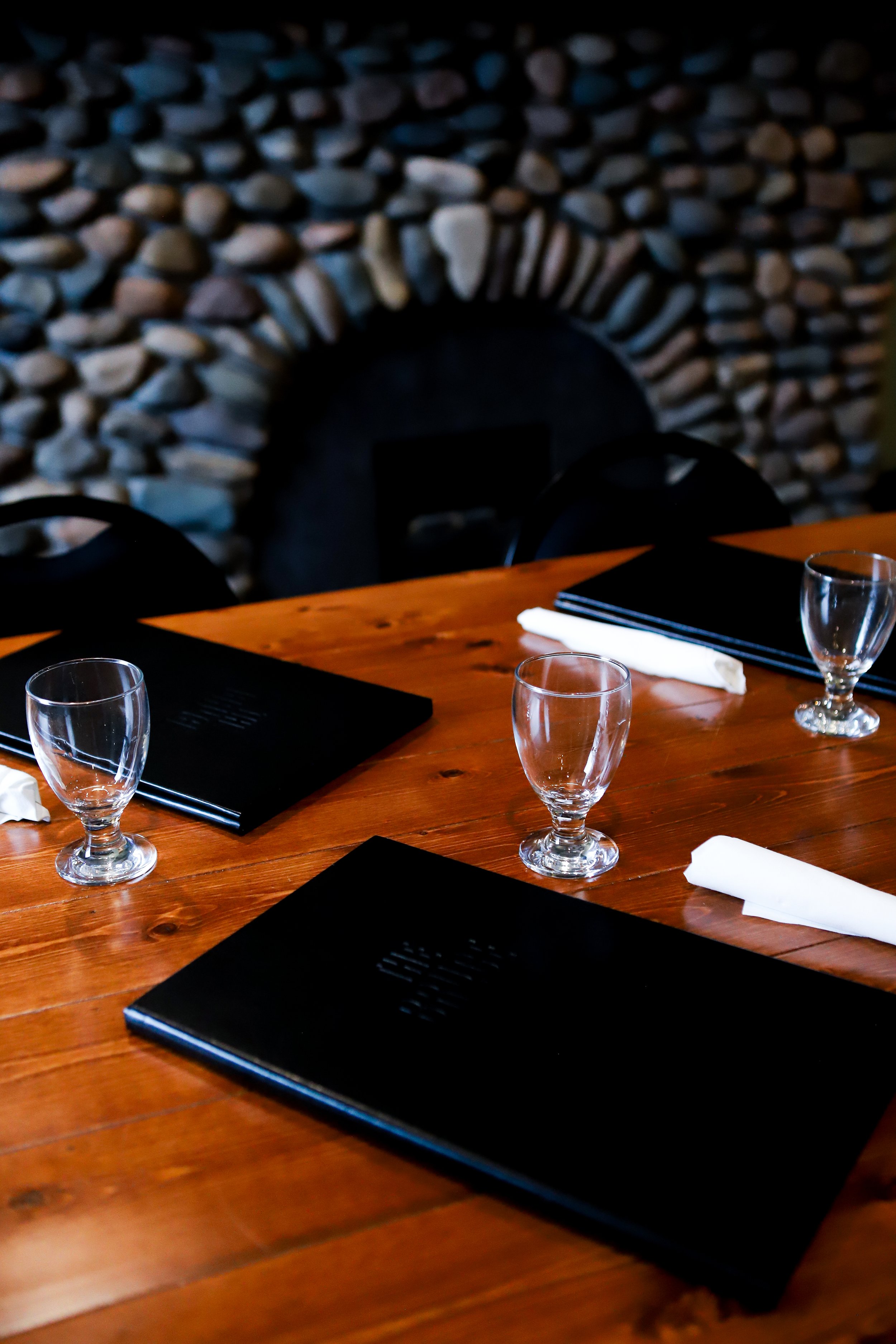The Mackenzie Room. Empty restaurant table with black menus, white napkins, wine glasses, and a stone fireplace in the background.