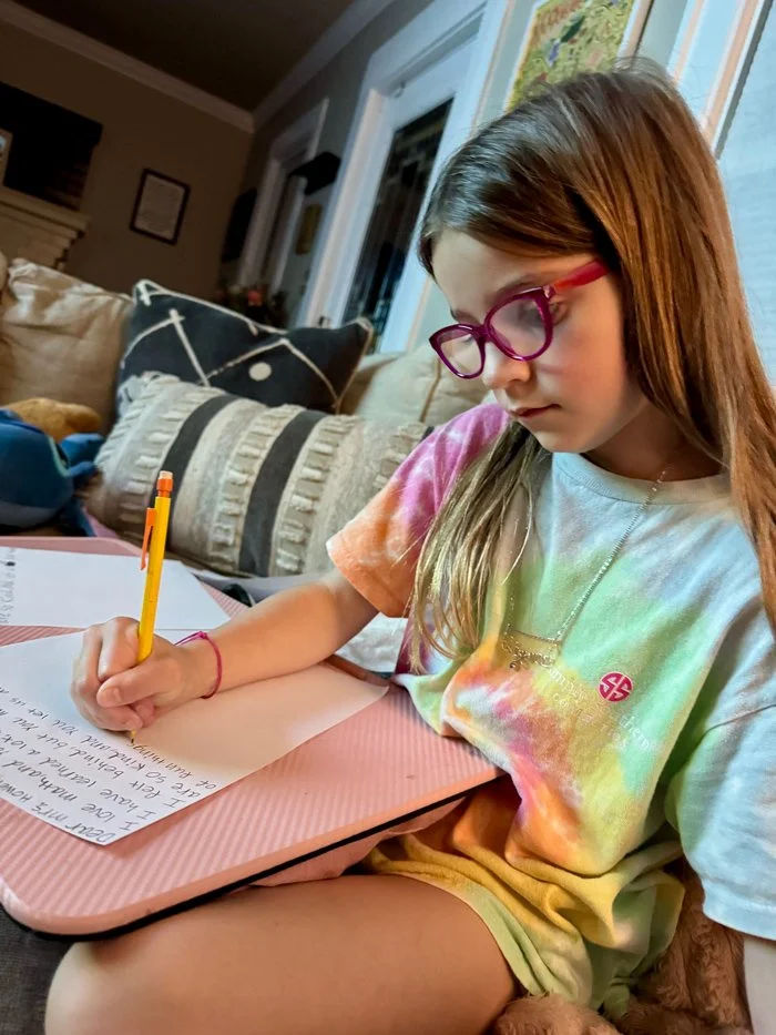 A young girl wearing purple glasses and a tie-dye shirt is sitting on a couch, writing on a piece of paper with a yellow pencil. There are pillows and other household items on the couch behind her.