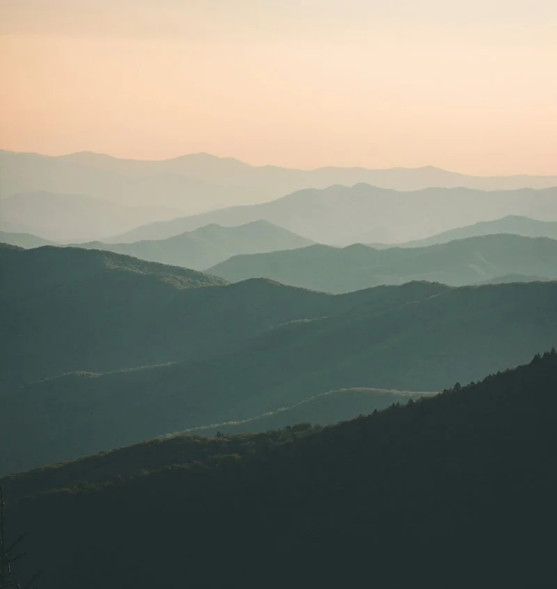 A scenic view of misty mountain ranges during sunset or sunrise with soft pastel colors in the sky.
