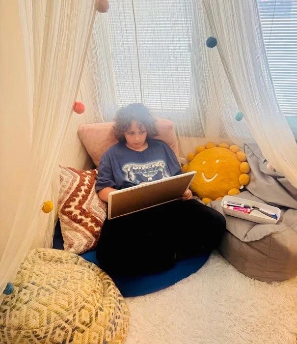 A child sitting on a blanket in a cozy, decorated nook, using a laptop. The space has pillows, a yellow smiley face pillow, and a tray with markers on a bean bag chair. She is surrounded by curtains and string lights with colorful pom-poms.