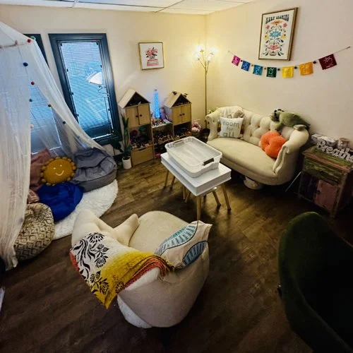 Living room with a white vintage sofa and a matching armchair, decorated with colorful pillows and a blanket, with a small table, a lamp, and decorative wall art, including a