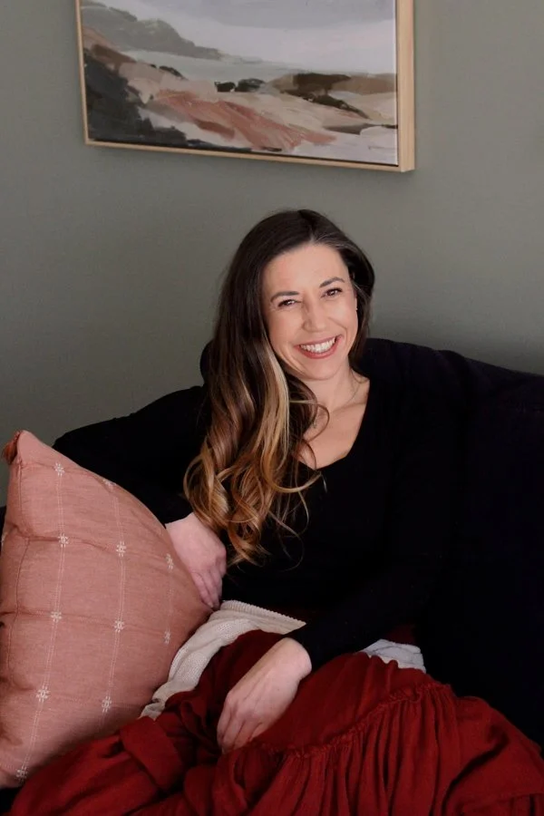 A woman with long brown hair, smiling, sitting on a black couch with a pink pillow, in a room with a painting of a landscape above her.