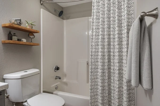 Small bathroom with a toilet, a shower with a patterned curtain, and two wall-mounted wooden shelves holding toiletries and a small plant.