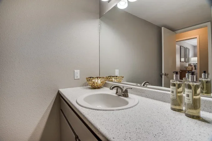 Bathroom vanity with a white speckled countertop, a white sink with silver faucet, soap bottles, and a mirror reflecting part of the room.