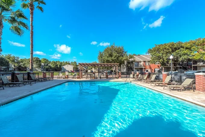 Outdoor swimming pool with clear blue water, surrounded by lounge chairs, trees, and a wooden pergola under a bright blue sky with a few clouds.