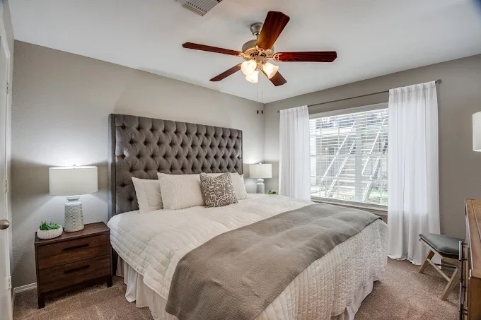 Bedroom with a large bed featuring a tufted gray headboard, white bedding, and multiple pillows. There are matching nightstands with lamps on either side of the bed. A ceiling fan with wooden blades is above. The room has a large window with sheer wh