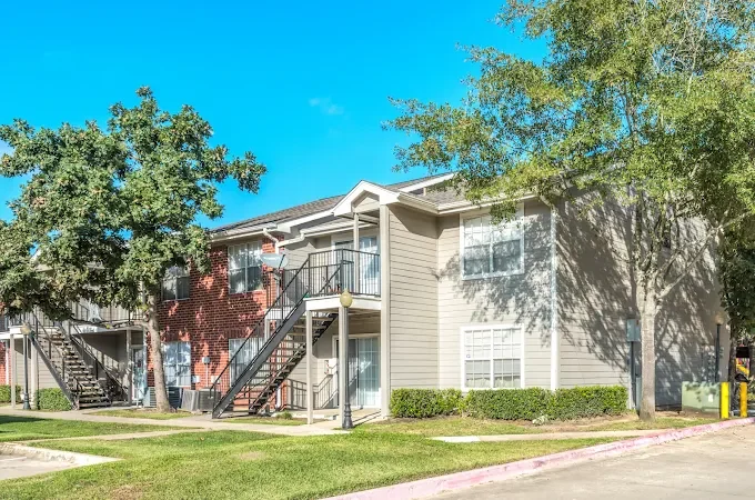 Exterior view of a two-story apartment building with outdoor staircases, surrounded by trees and a well-maintained lawn in a residential area.