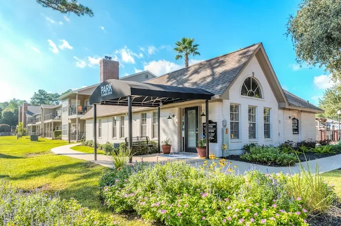 Exterior view of a white residential building with a sloped roof and large arched window, surrounded by a well-maintained lawn and garden, with a black awning over the entrance that says 'PARK', and a clear blue sky overhead.