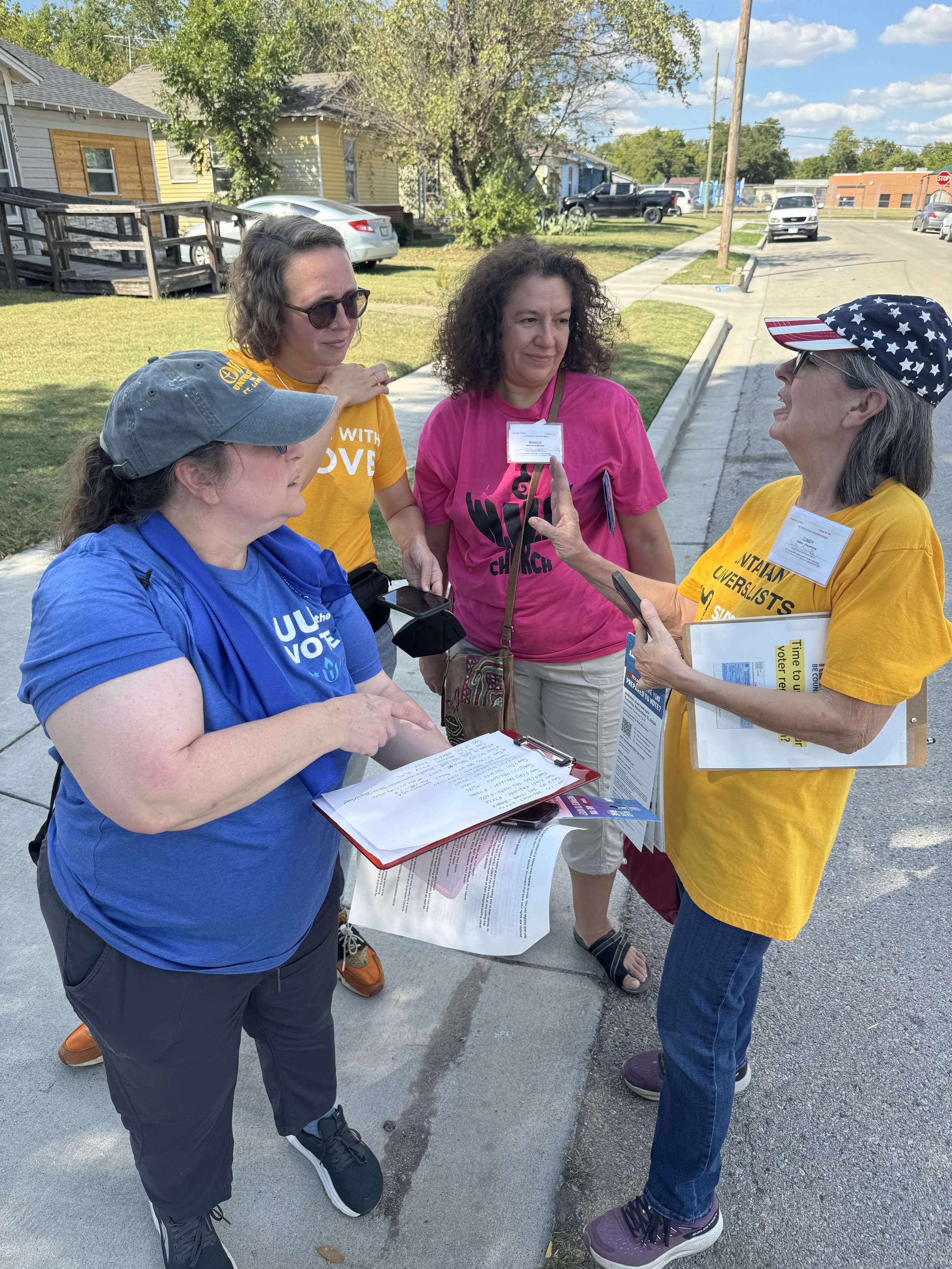 Group of women engaged in conversation outdoors on a sunny day, some wearing colorful t-shirts and holding clipboards or documents.