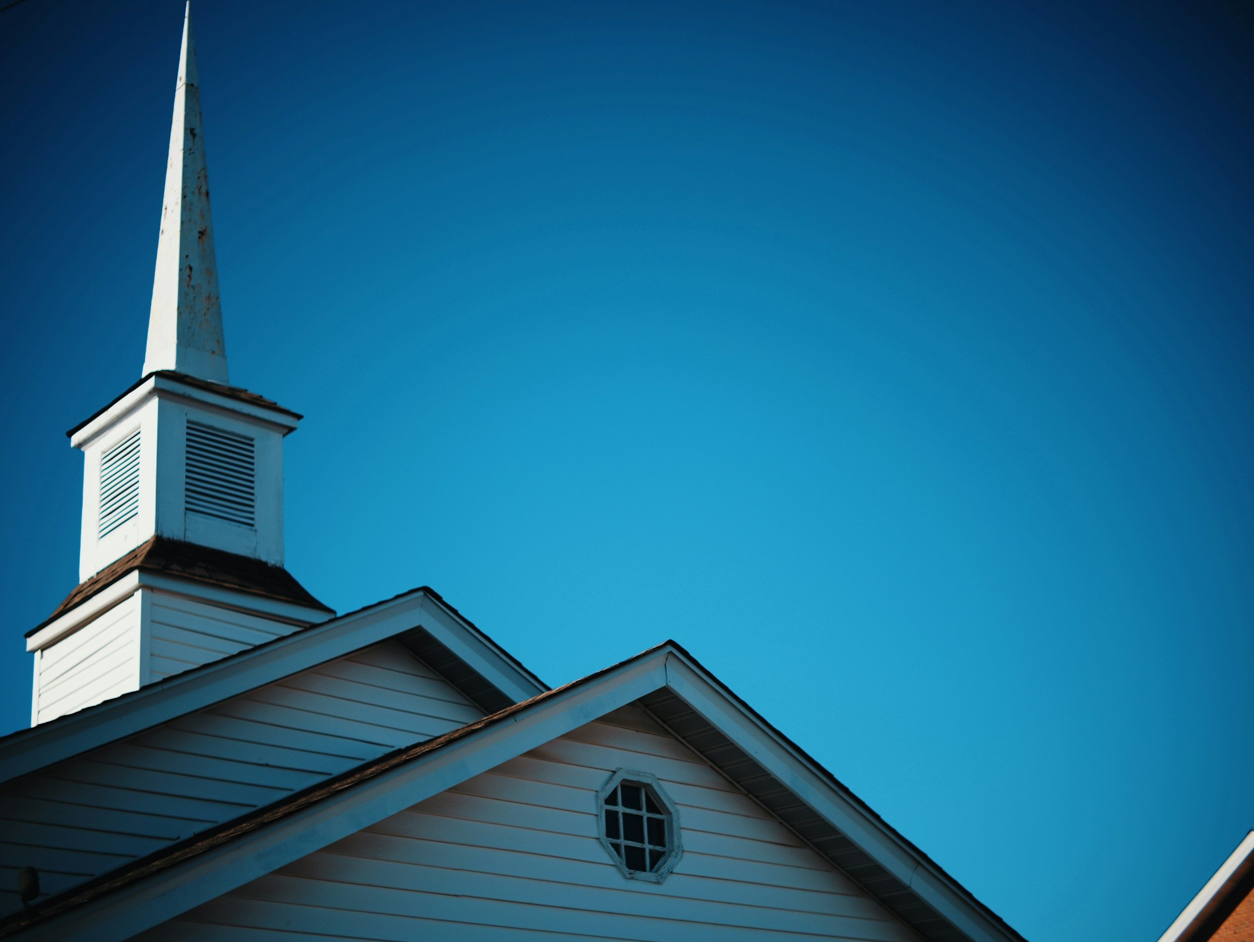 A white church steeple with a pointed tip and a rectangular base against a clear blue sky, attached to a white building with a shingled roof.