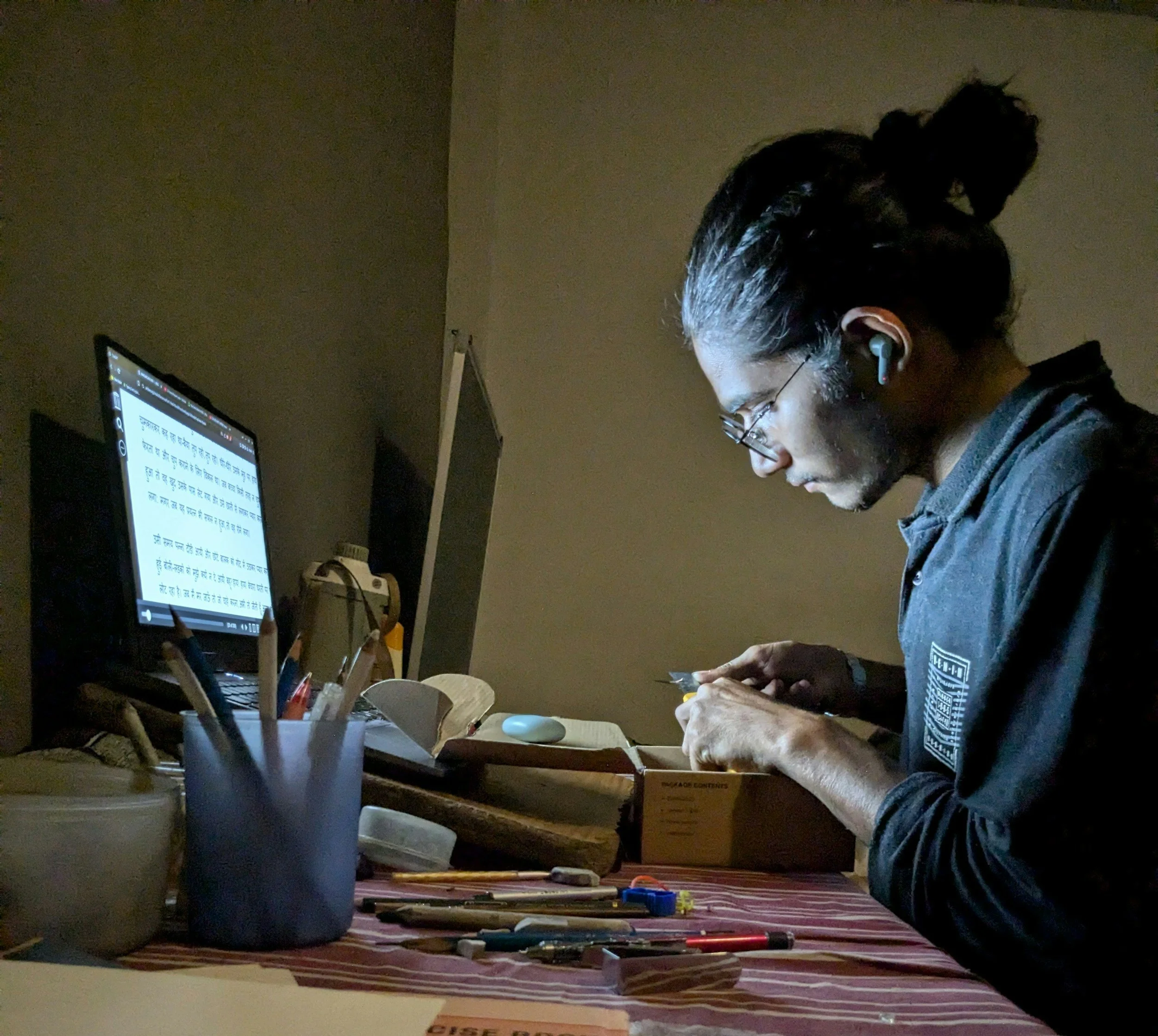 A person wearing glasses and airpods, working at a desk, illuminated by a computer screen displaying text in an Asian language. The desk holds pens, markers, and a blue trash can, with a striped rug on the floor.
