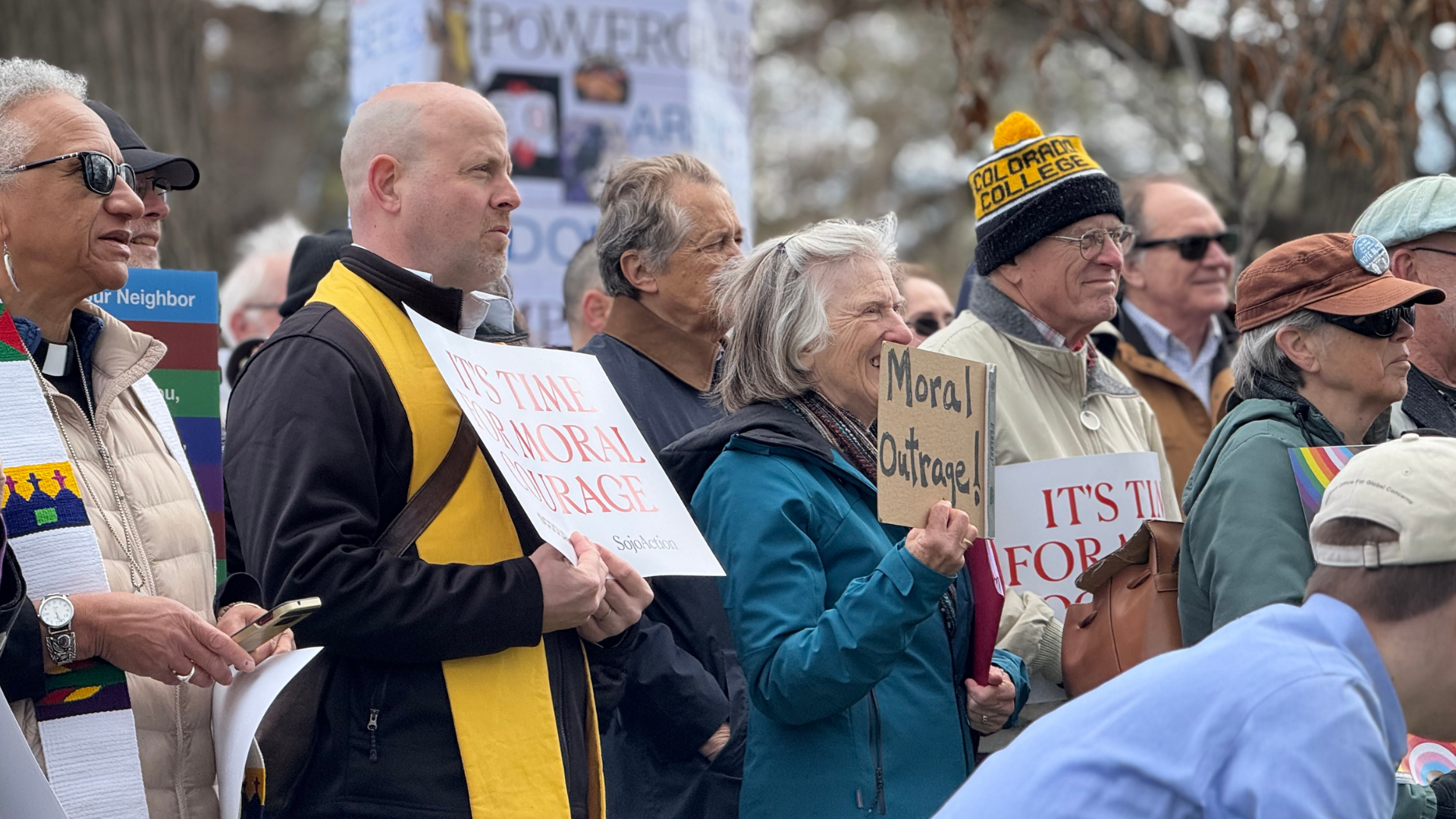 A group of diverse people participating in a protest or rally outdoors, holding signs with messages like "It's time for moral courage" and "Moral outrage." Some individuals are wearing hats and sunglasses, and the crowd appears attentive and engaged.