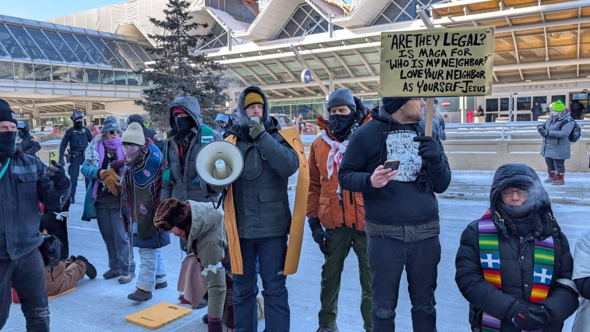 A group of people dressed warmly in winter clothing participating in an outdoor protest or demonstration. One person holds a sign with a quote about legal rights and love thy neighbor. Others are holding phones, a megaphone, and are visibly engaged. The background features a modern building, a tree, and other pedestrians.
