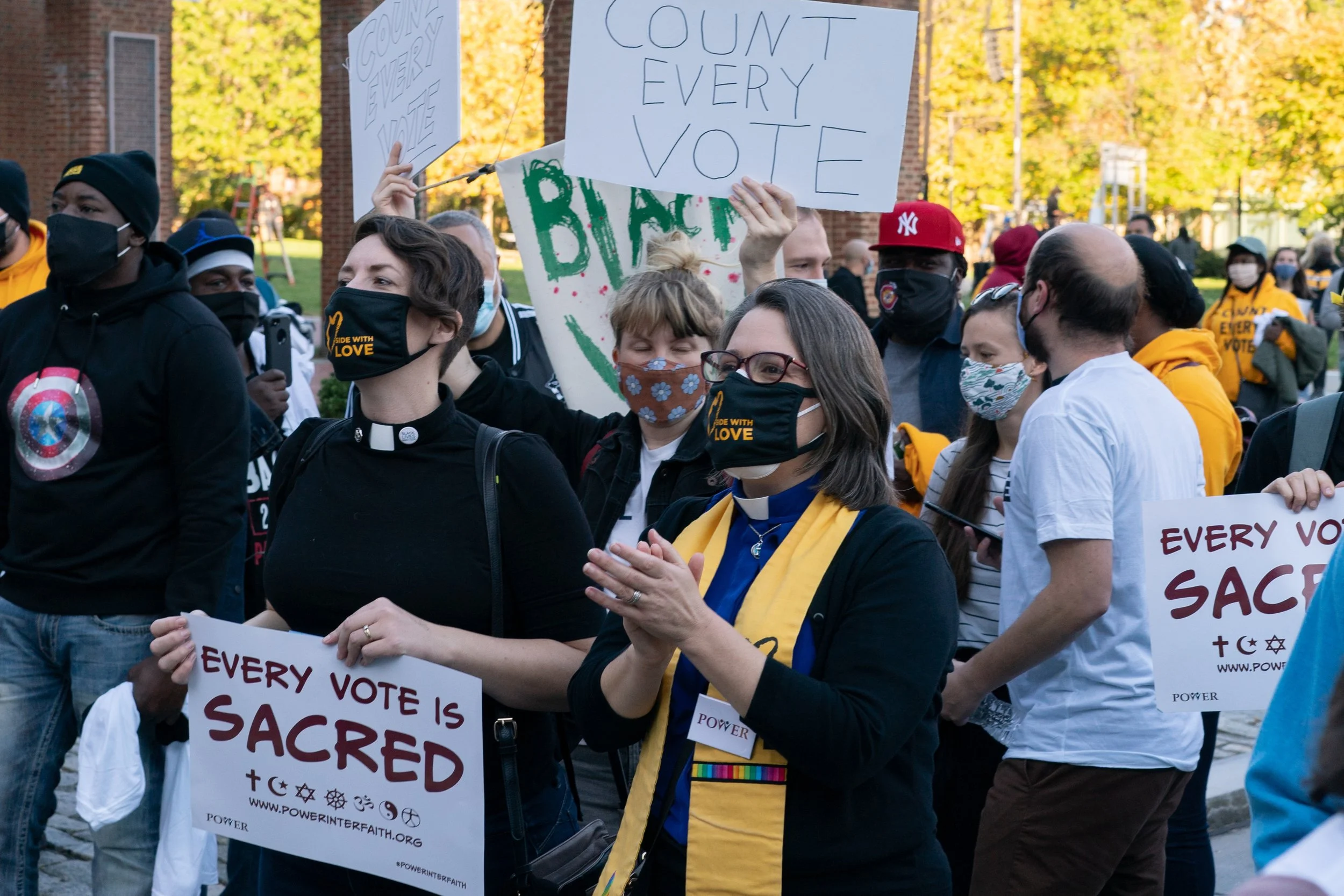 Group of diverse people participating in a protest or rally, holding signs that promote voting, including one that says 'Count Every Vote' and another that says 'Every Vote is Sacred.' The participants are wearing masks and appear to be advocating for voting rights and justice.