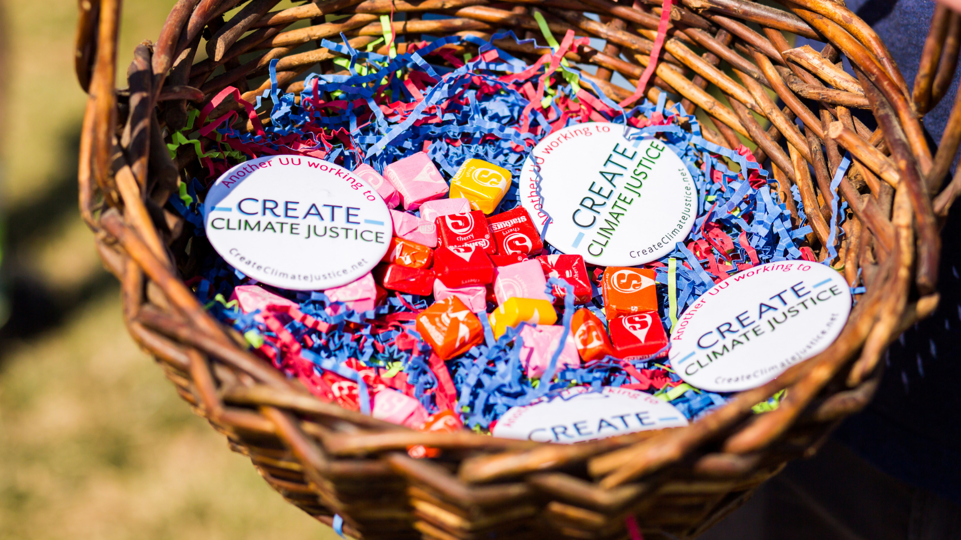 A wicker basket filled with colorful shredded paper, buttons that say 'S', and stickers with the words 'Create Climate Justice' on a white background with pink and green text.