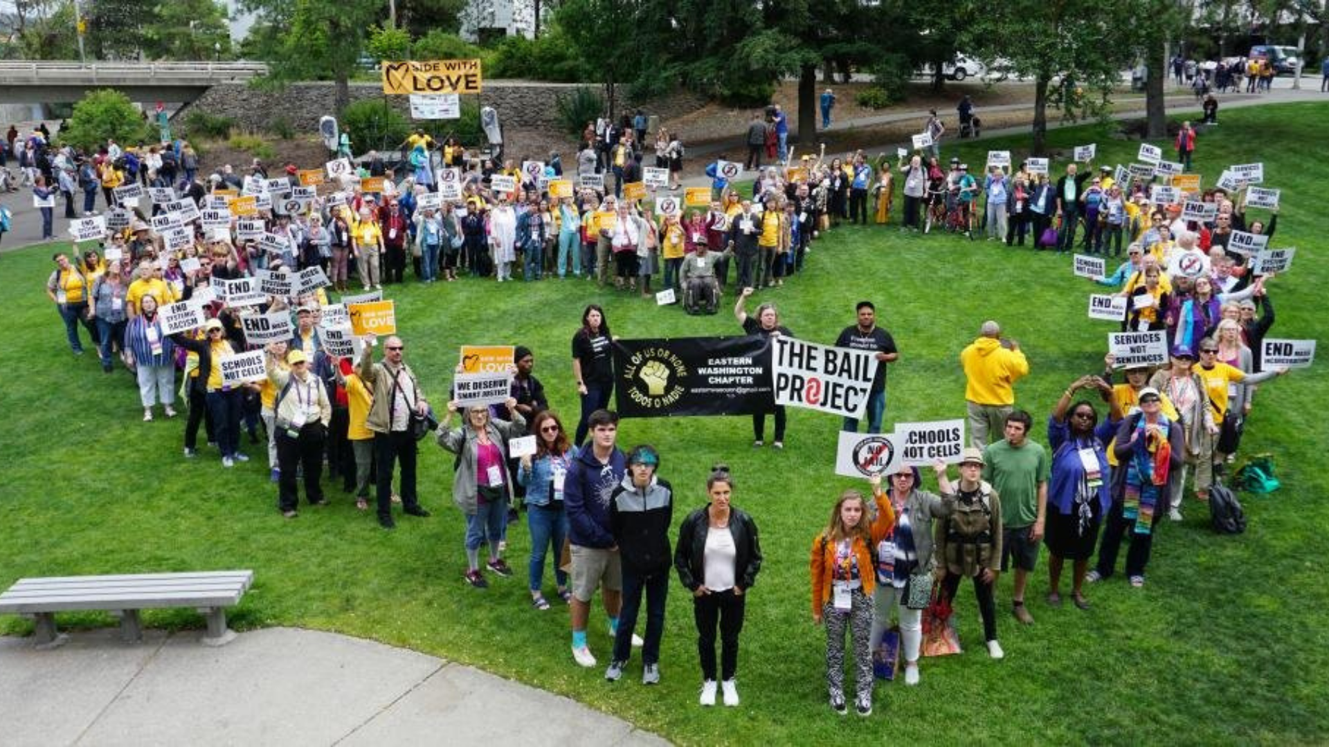 A large group of people participating in a protest or rally in a park, holding signs advocating for education and schools without cells, with banners and signs in support of love and ending cell phone use in schools.