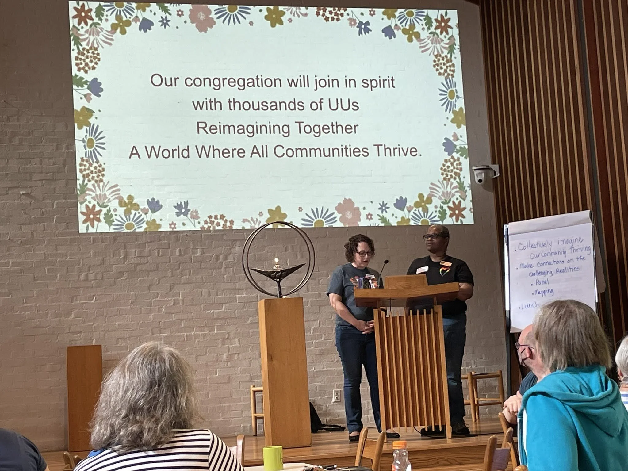 A projected slide with a floral border and the message: 'Our congregation will join in spirit with thousands of UUs Reimagining Together A World Where All Communities Thrive.' Two people stand at a wooden podium, speaking or presenting to an audience in a room with brick walls and a wooden panel on the right. Several audience members are visible, including a child in a turquoise hoodie and adults, some with gray or curly hair.