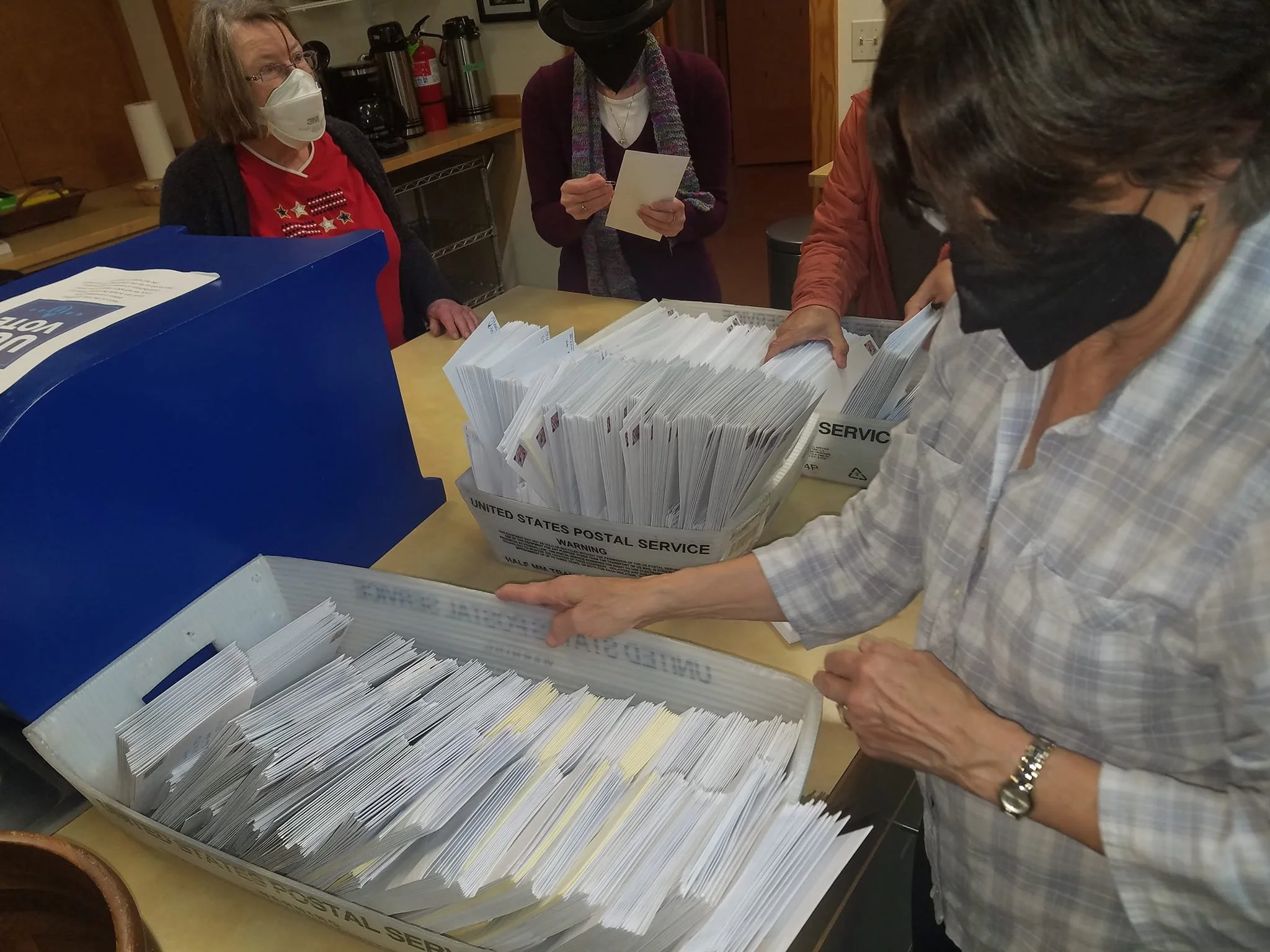 Multiple people wearing masks sorting and organizing envelopes at a table, possibly at a post office or mailing center.