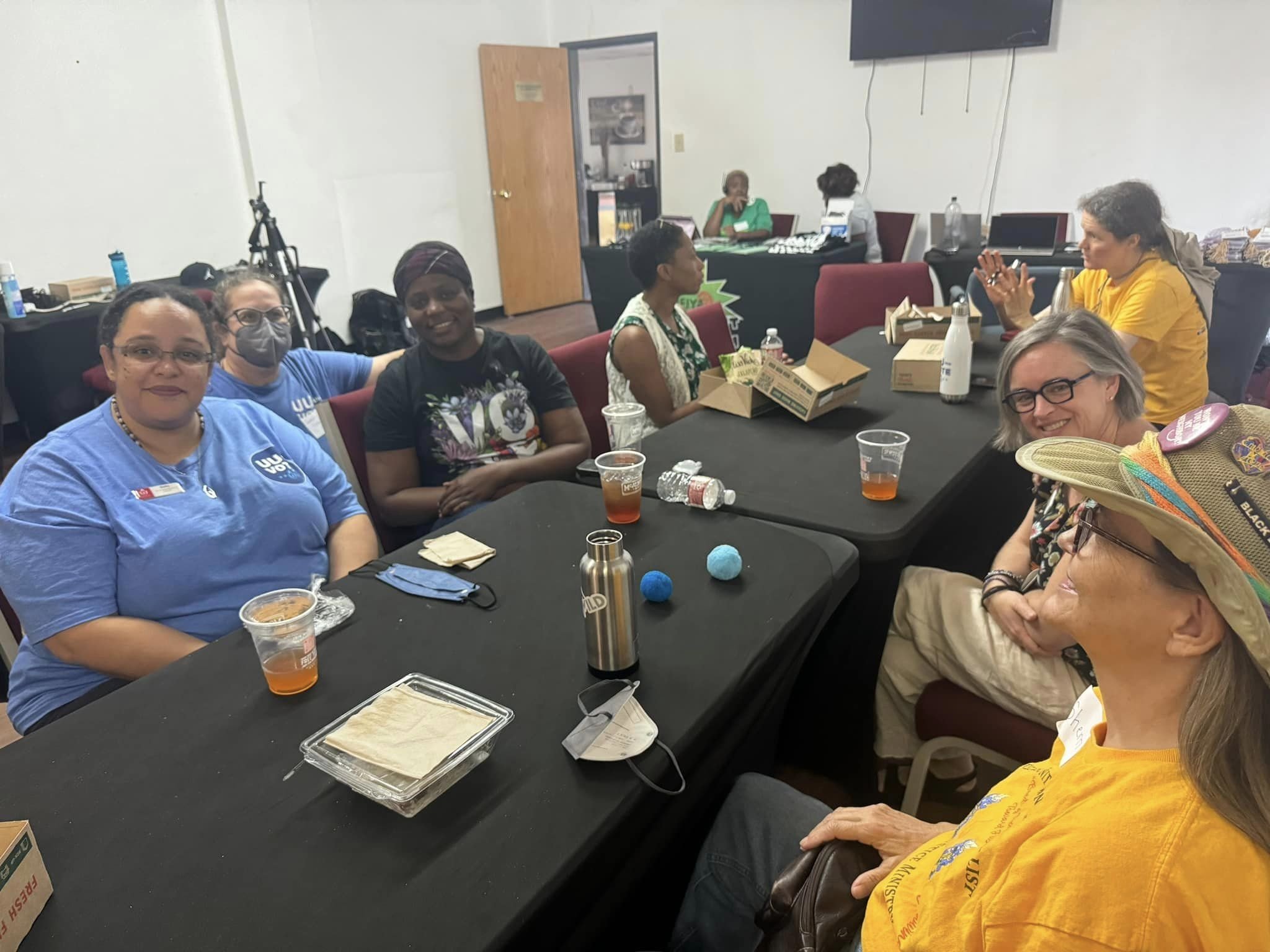 Group of diverse women sitting around a black conference table in a meeting room, with some women smiling and some talking. There are snacks, drinks, a water bottle, and miscellaneous items on the table.